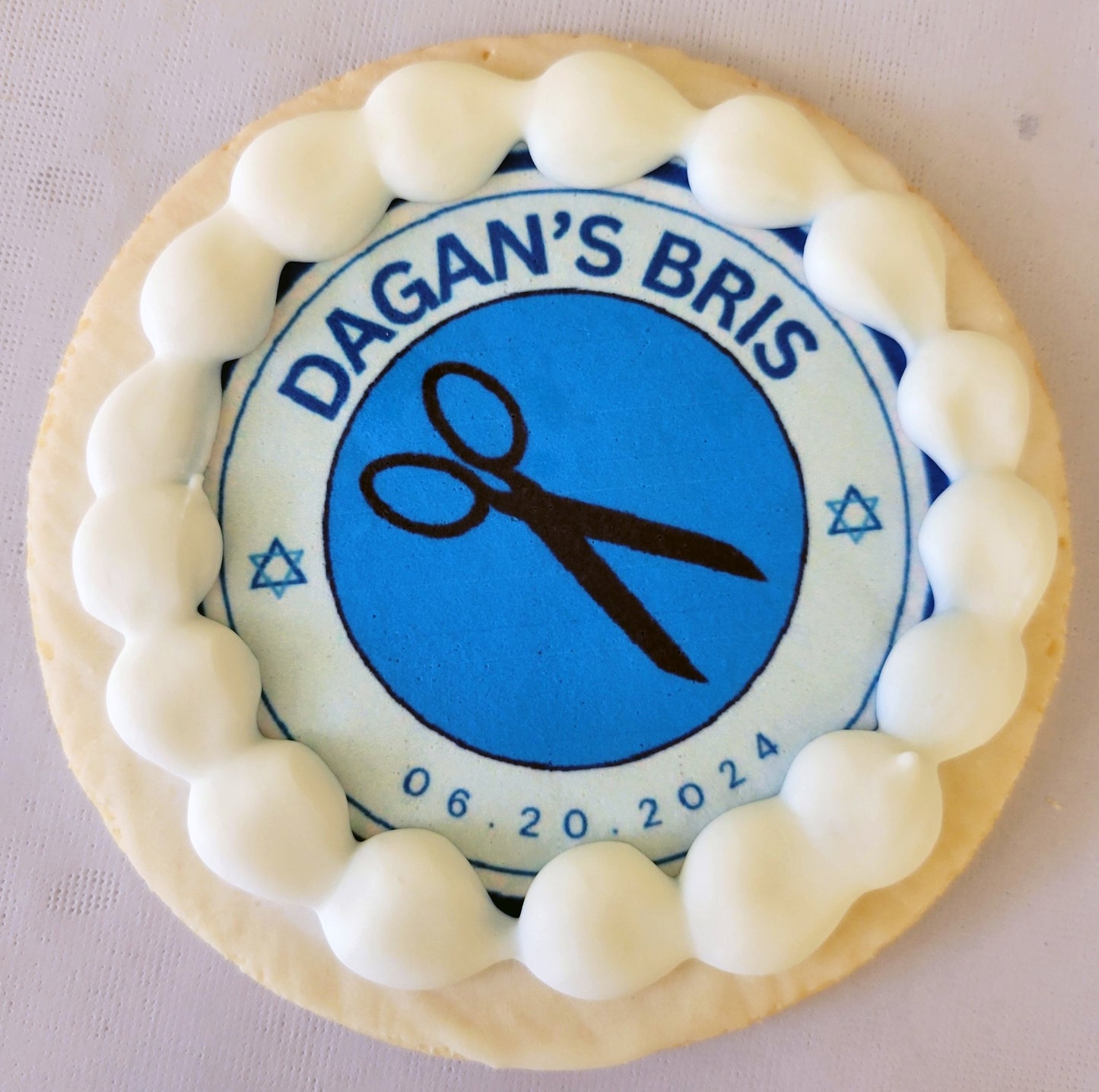 Cookie with blue circle featuring 'Dagan's Bris' and scissors, surrounded by white icing on a white background.