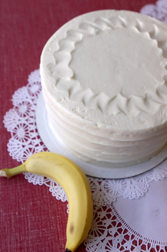 A frosted banana cake on a plate with a banana to the side, presented on a lace tablecloth.