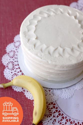 A frosted banana cake on a plate with a banana to the side, on a lace tablecloth, with a shipping available icon.