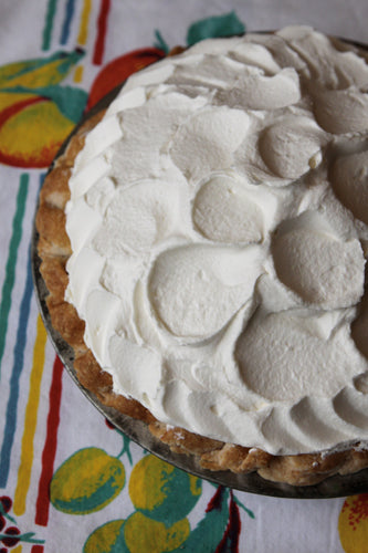 A freshly made banana cream pie with whipped cream on top, placed on a table with a colorful fruit-patterned tablecloth.