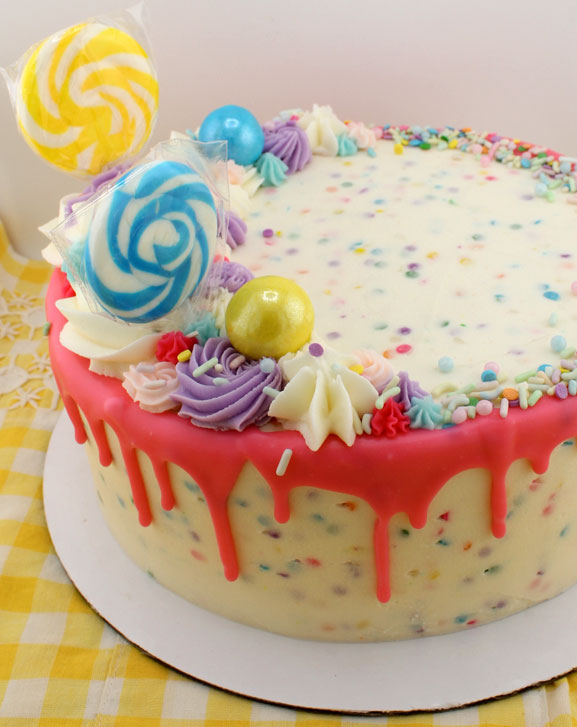 A colorful 'Sweet Celebration' cake with white chocolate drip, multicolored sprinkles, and decorative candies on top, displayed on a yellow checkered background.