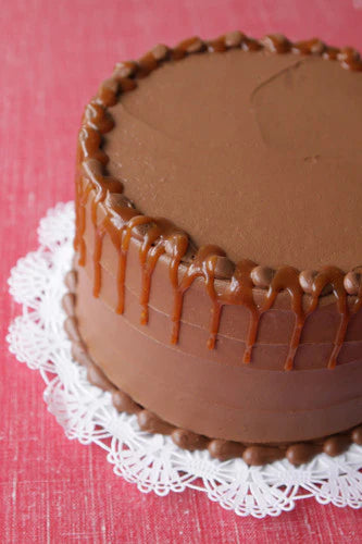 A chocolate cake with smooth chocolate frosting, topped with a decorative border of chocolate candies and a luscious caramel drizzle dripping down the sides, presented on a white lace doily.