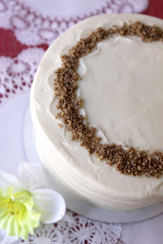 A round carrot cake with cream cheese frosting garnished with chopped pecans, and served on a white plate with a red and white lace background.