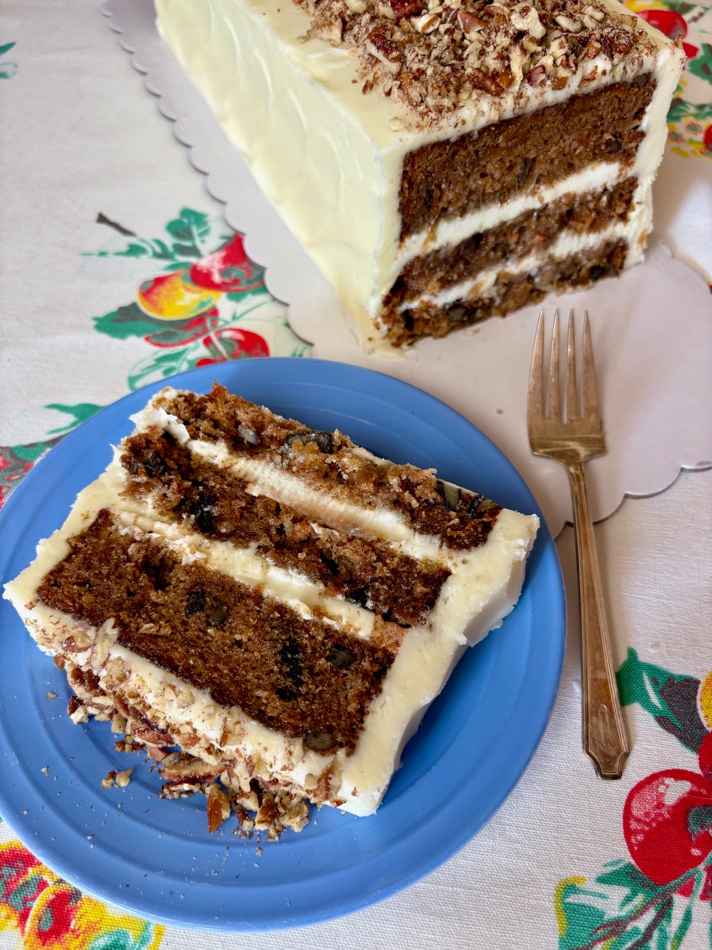 Slice of layered carrot cake on a blue plate with a fork, on a colorful tablecloth.