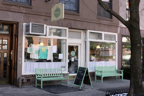 Exterior view of Billy's Bakery in Chelsea, New York City, featuring a brownstone facade, large display windows with merchandise, two green benches, and a chalkboard sign on the sidewalk.