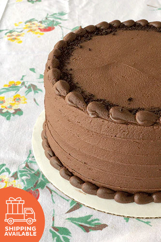 A round chocolate cake with buttercream frosting, placed on a plate, with a shipping available icon in the bottom right corner.