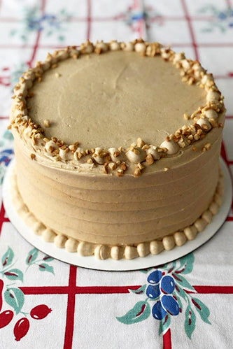 A frosted chocolate peanut butter cake with peanut crumbles garnished around the border, served on a plate with a patterned tablecloth underneath.