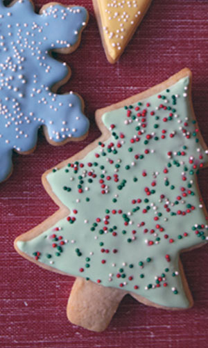 A sugar cookie cut into a Christmas tree shape, decorated with green, red, and white icing and sprinkles on a red background.