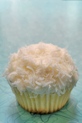 A coconut cupcake topped with vanilla buttercream and fluffed with coconut flakes, on a blue patterned surface.