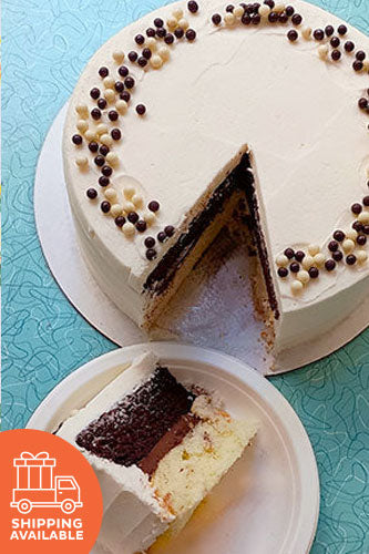 A chocolate and vanilla layer cake with buttercream frosting, decorated with chocolate pearls, with one slice removed, placed on a plate, with a shipping icon overlay at the bottom right corner.