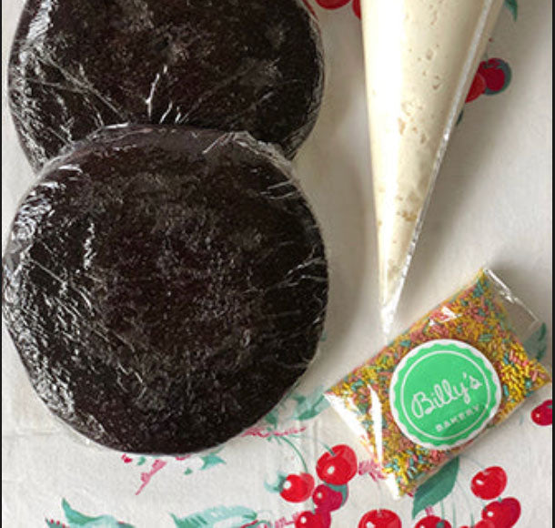 Two round chocolate cakes wrapped in plastic, a piping bag of white buttercream frosting, and a pouch of multicolored sprinkles with a branded Billy's Bakery spatula, all presented on a paper with a Christmas pattern.