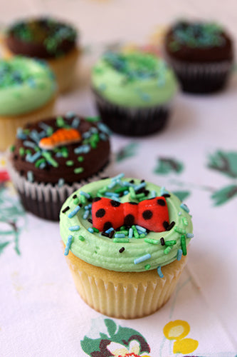 Cupcakes with green frosting and ladybug decorations on a floral tablecloth.