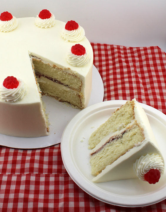 A round lemon raspberry cake with white frosting and red raspberry garnishes on top. Slice of cake placed on a plate showing the inside filling of raspberry jam and vanilla buttercream.