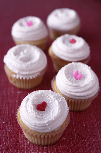 A group of cupcakes with white frosting and heart-shaped decorations, presented on a red surface.