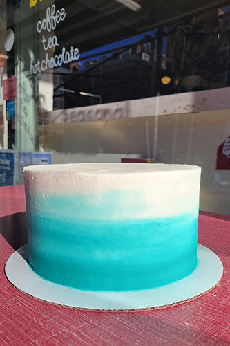 A beautifully decorated Ombre Parfait cake with a gradient of light to dark blue, served on a table with a red surface, in front of a counter with signage offering coffee, tea and hot chocolate.