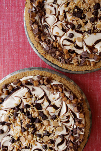 Two peanut butter chocolate pies with graham cracker crusts, topped with chocolate syrup and peanut butter cups, displayed on a red surface.