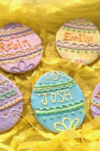 Decorated egg-shaped sugar cookies with royal icing, displayed in a set of two. The cookies are adorned with pastel colors and have names written on them.