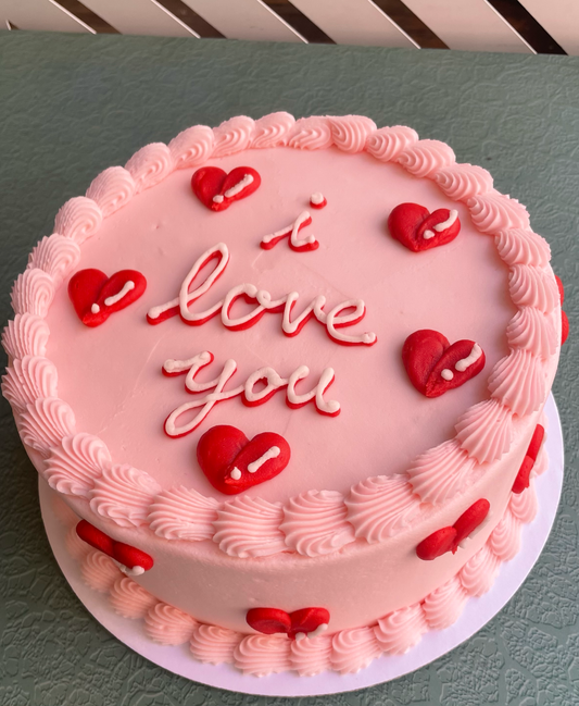 A frosted cake with pastel pink buttercream and decorated with puffy red bubble hearts and the words 'i love you' in cursive on top.