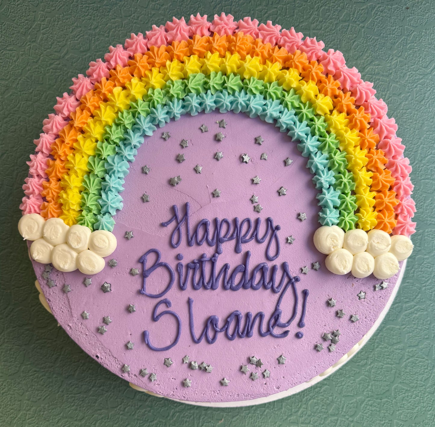 A beautifully decorated birthday cake with a rainbow-shaped top, pastel colors, and the message 'Happy Birthday' in the center.