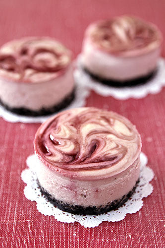 Three small round raspberry cheesecakes with a chocolate crust on a red surface, garnished with raspberries on top.
