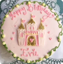 A round pink birthday cake decorated with a pink and gold castle in the center, "Happy Birthday" written in pink icing at the top, and "Evie" written in pink icing at the bottom. The cake also features green vine-like borders and small yellow and pink accents around the edges.