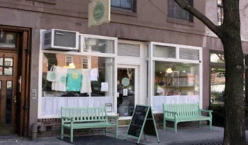 Exterior of Billy's Bakery in Chelsea, NYC, featuring a storefront with large windows displaying baked goods, two green benches, and a chalkboard sign on the sidewalk.