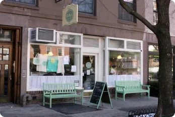 Exterior view of Billy's Bakery in New York City, featuring a brown building with large windows displaying baked goods, two green benches, a sidewalk sign, and a tree on the right.