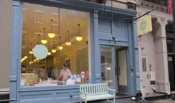 Exterior of Billy's Bakery in Tribeca, New York City, featuring a large storefront window displaying baked goods and a person inside, with a white bench outside and blue-painted facade.