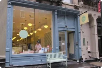 Exterior view of Billy's Bakery storefront in New York City with a person inside, a light blue facade, large windows, and a white bench outside.