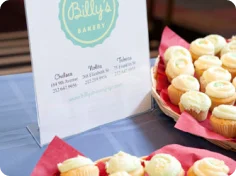 A menu board from Billy's Bakery advertising cupcakes and other baked goods, positioned next to a basket filled with small, frosted cupcakes.
