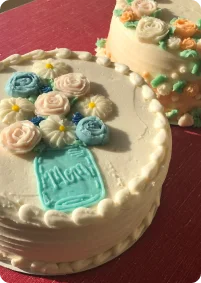 A white frosted cake decorated with a light blue mason jar design filled with pink, blue, and white icing flowers. In the background, another cake with floral decorations is partially visible.