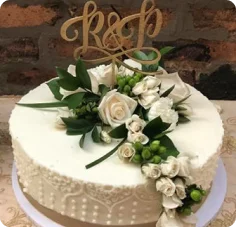 A white, single-tier wedding cake decorated with white roses, green foliage, and a gold "R & P" cake topper, set against a blurred brick background.