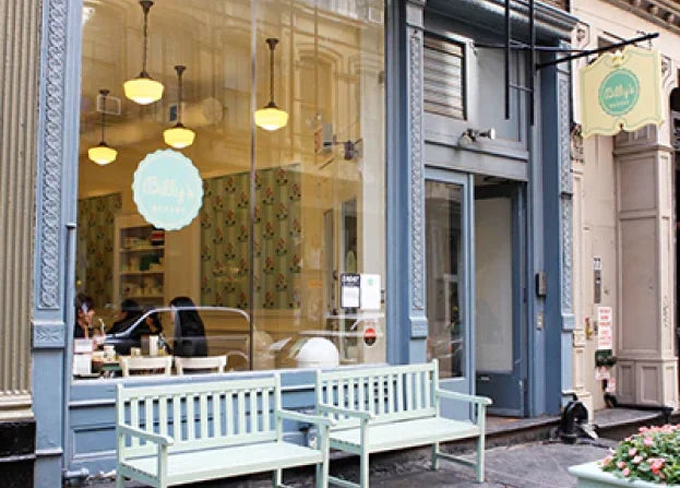 Exterior view of Billy's Bakery in Tribeca, New York City, featuring its blue storefront, large windows displaying the interior, two white benches outside, and the bakery's sign.