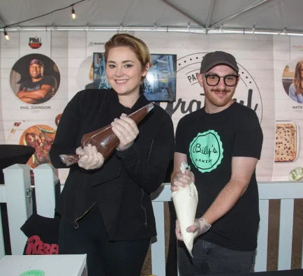 Two smiling bakers, a woman and a man, holding piping bags in a bakery setting with a 'Billy's Bakery' logo on the man's shirt.