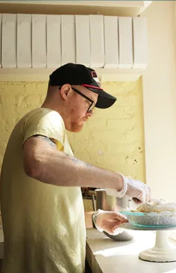 A man in a cap and glasses wearing gloves prepares food on a counter, with white boxes stacked above him.