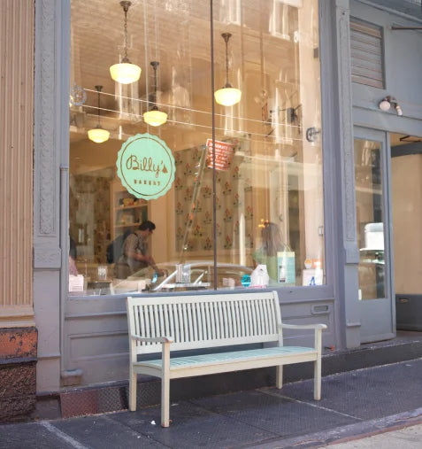Exterior of Billy's Bakery in New York City, featuring a light-colored wooden bench in front of the large glass window displaying the 'Billy's Bakery' logo and interior view.