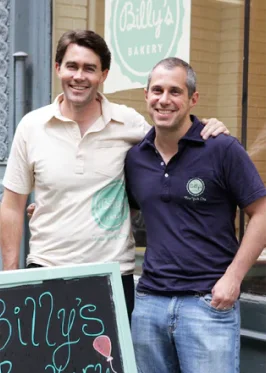 Two smiling men, one in a beige Billy's Bakery polo and one in a purple Billy's Bakery polo, standing outside Billy's Bakery.