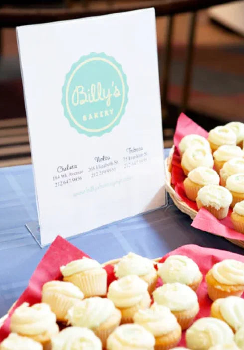 Assortment of vanilla cupcakes from Billy's Bakery displayed on a table with a sign featuring the bakery's logo and New York City locations.