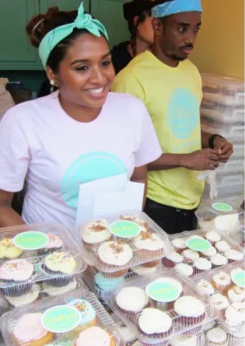 Two smiling people, a woman in a mint green headband and a man in a blue headband, stand behind a display of many clear containers filled with various cupcakes at what appears to be a bakery or market stall. The woman is wearing a light pink t-shirt with a mint green circle design, and the man is wearing a yellow t-shirt with a similar design.