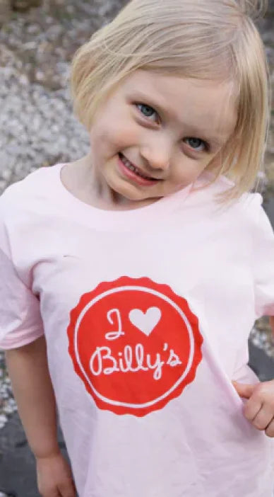 Young blonde girl smiling and looking at the camera, wearing a light pink t-shirt with a red circular logo that says "I heart Billy's" in white script.
