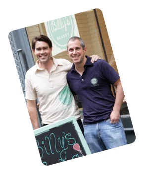 Two smiling men, one in a beige Billy's Bakery polo and one in a purple Billy's Bakery polo, standing outside Billy's Bakery.
