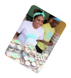A smiling woman in a light-colored shirt and a man in a yellow shirt stand behind a counter filled with baked goods.