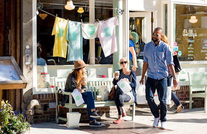 People enjoying coffee with shopping bags outside Billy's Bakery, with colorful t-shirts hanging on a line in front of the shop window.