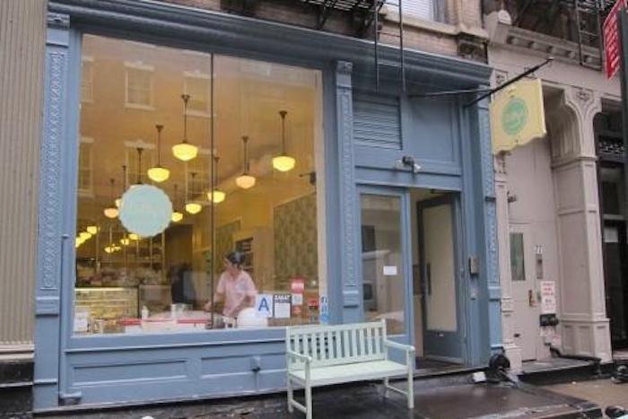 Exterior of Billy's Bakery in Tribeca, NYC, showing the blue storefront, large windows displaying baked goods and a person inside, with a light green bench outside.