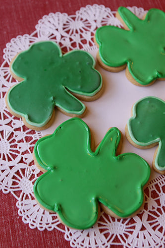 This vibrant image showcases several homemade, shamrock-shaped sugar cookies, each generously coated with bright green royal icing. A subtle, lighter green frosting outlines the edges of each cookie, adding a delicate detail. The cookies are artfully arranged on a round, white lace paper doily, which provides a beautiful textural contrast. The doily and cookies are set against a soft, reddish or maroon-colored surface, possibly a tablecloth, which makes the green of the shamrocks pop. The lighting appears s