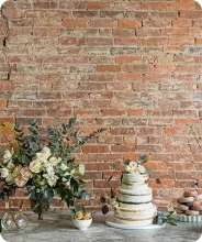 Elegant wedding cake with white and gold tiers, surrounded by white floral arrangements and desserts on a table with a brick wall backdrop.