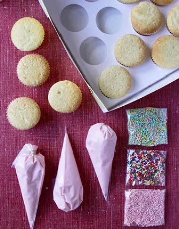 A flat lay image showing a cupcake decorating kit. The kit includes several plain vanilla cupcakes in a white box and scattered on a pink surface, three piping bags filled with pink frosting, and three bags of colorful sprinkles and edible decorations.