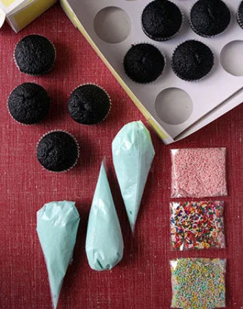 Overhead view of a cupcake decorating DIY kit on a red surface, featuring six baked chocolate cupcakes, three piping bags filled with light blue frosting, and three clear bags of assorted sprinkles in different colors and shapes. A white cupcake box with empty compartments and some cupcakes is visible in the background.