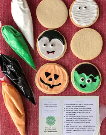 Overhead shot of Halloween-themed sugar cookies and decorating supplies. Four piping bags filled with white, green, black, and orange royal icing are on the left. On the right, decorated cookies include a mummy, a vampire, a jack-o'-lantern, and Frankenstein's monster, alongside two plain round cookies. A printed instruction sheet for decorating is at the bottom right.