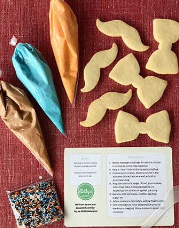 A DIY cookie decorating kit featuring sugar cookies shaped like mustaches and bow ties, three piping bags filled with frosting in blue, orange, and brown, and a bag of colorful sprinkles, laid out on a red surface. Instructions for decorating are also visible.